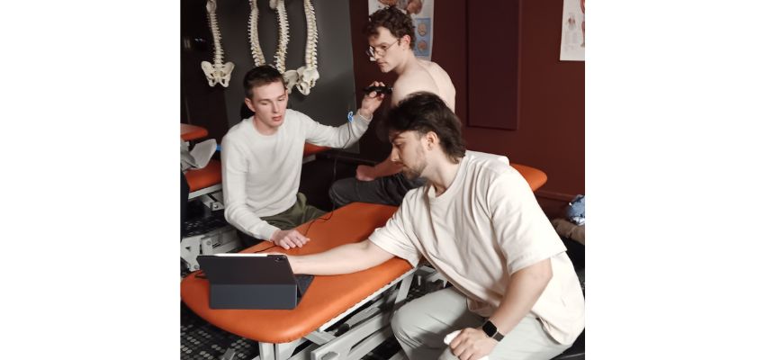 Students practice a clinical procedure using monitoring equipment while gathered around a patient on an exam table.