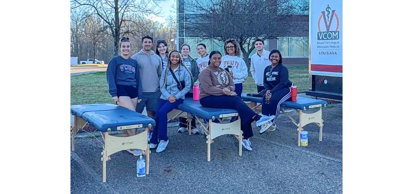 A group of students pose outdoors around portable treatment tables set up near a VCOM Louisiana mobile unit.