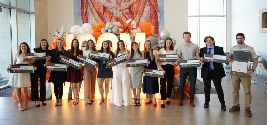 A group of students stand in a line holding large ceremonial checks at a formal event with decorative balloons behind them.