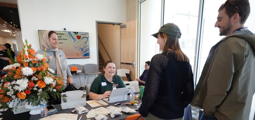 Staff members greet attendees at a table with informational materials and flowers during a campus event.