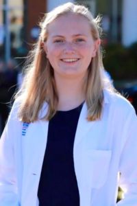 A medical student in a white coat smiles in a professional outdoor headshot.