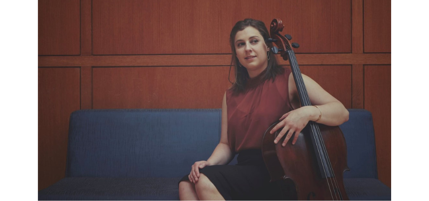 A woman sits upright on a bench in front of a wood paneled interior, holding her cello.