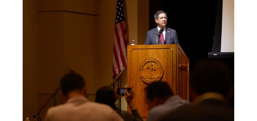 A speaker stands at a podium delivering remarks during a formal event with an audience seated in front.