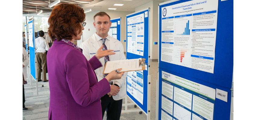 A presenter reviews a research poster with an attendee while holding a clipboard at a conference.