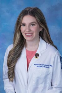 A student physician in a white coat smiles in a professional headshot against a blue background.