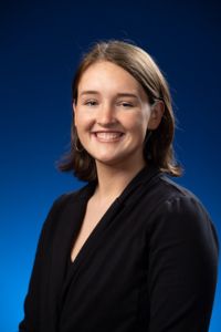 A woman in a black blazer smiles in a studio portrait with a blue gradient background.