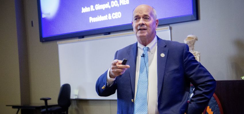 A man in a suit speaks while holding a remote during a presentation with a projected slide behind him.