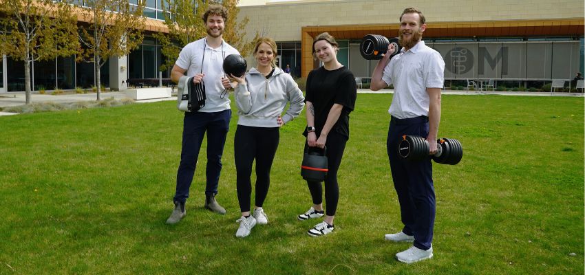 Four people stand on a lawn outside a campus building holding dumbbells and a medicine ball, smiling at the camera.