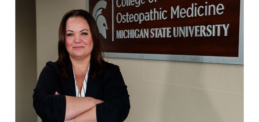 A woman stands with arms crossed in front of a wall sign reading “College of Osteopathic Medicine, Michigan State University.”