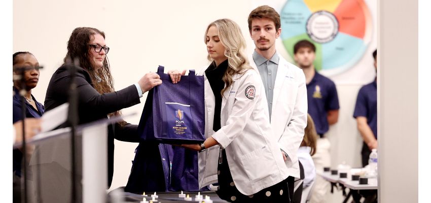 A student in a white coat receives a bag during a ceremony while others stand nearby in a classroom setting.