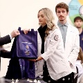 Astudentinawhitecoatreceivesabagduringaceremonywhileothersstandnearbyinaclassroomsetting