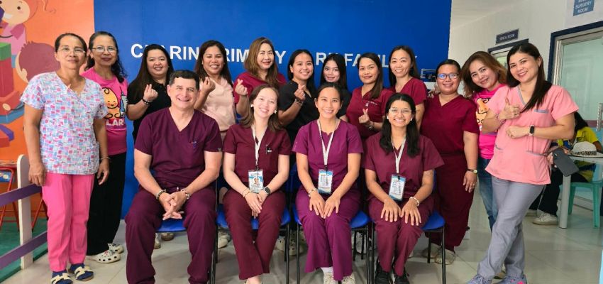 A group of healthcare workers and students in scrubs pose together in a clinic room, many making hand gestures and smiling.