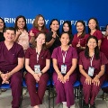 Agroupofhealthcareworkersandstudentsinscrubsposetogetherinaclinicroommanymakinghandgesturesandsmiling