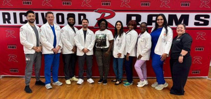 A group of students and faculty in white coats and scrubs stand in a gymnasium in front of a red “Rams” backdrop.