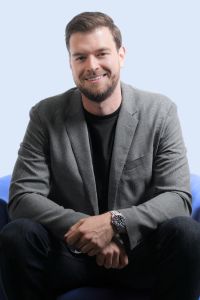 A man in a gray blazer and black shirt sits on a chair, smiling in a studio portrait with a light background.