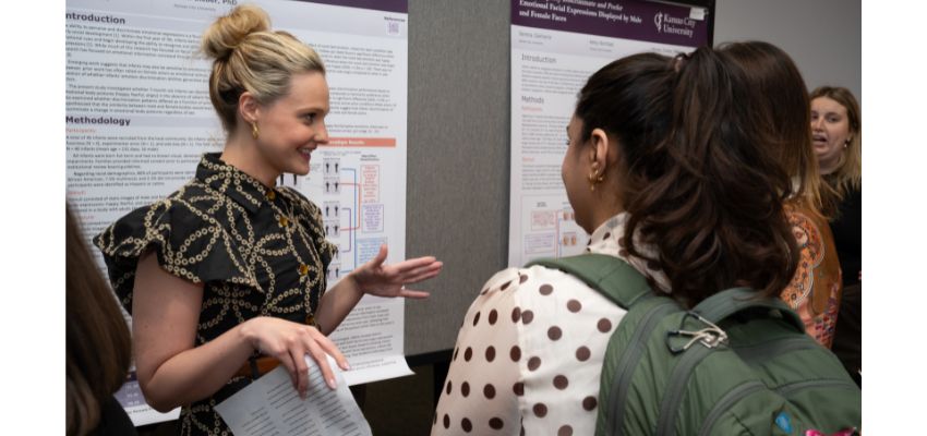 A woman presents a research poster while speaking with an attendee wearing a backpack at an academic conference.