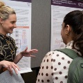 Awomanpresentsaresearchposterwhilespeakingwithanattendeewearingabackpackatanacademicconference