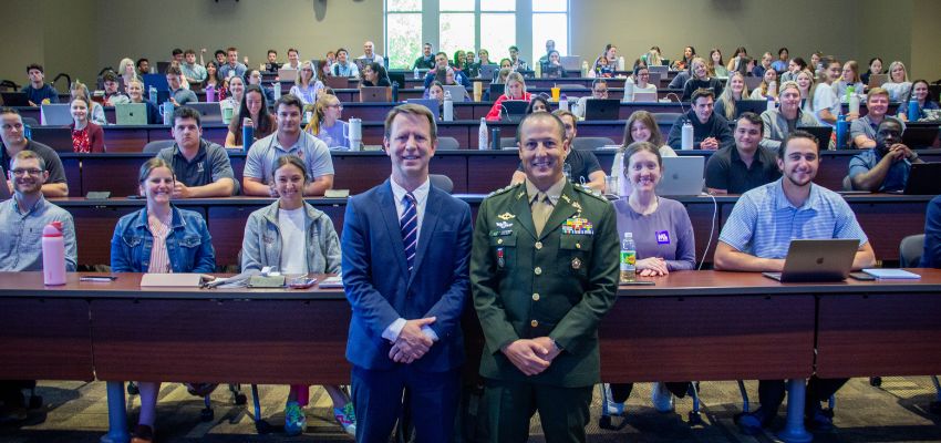 Two men, one in a suit and one in a military uniform, stand at the front of a large lecture hall filled with seated students.