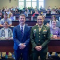 Twomenoneinasuitandoneinamilitaryuniformstandatthefrontofalargelecturehallfilledwithseatedstudents