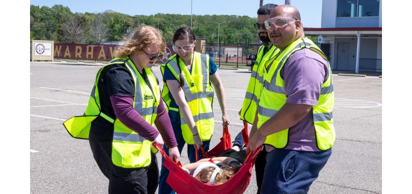 Three individuals wearing high-visibility vests carry a person on a stretcher during an outdoor emergency response training.