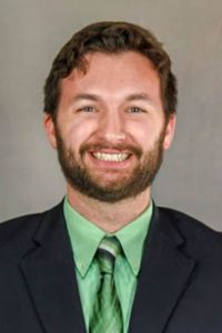 A man with a beard smiles in a professional headshot wearing a suit with a green shirt and tie against a neutral background.