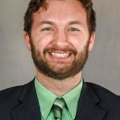 Amanwithabeardsmilesinaprofessionalheadshotwearingasuitwithagreenshirtandtieagainstaneutralbackground