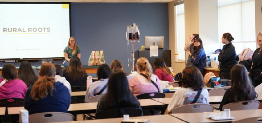 A classroom of students listens to a presenter standing near a screen displaying “Rural Roots,” with medical training equipment visible.