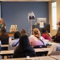 AclassroomofstudentslistenstoapresenterstandingnearascreendisplayingRuralRootswithmedicaltrainingequipmentvisible