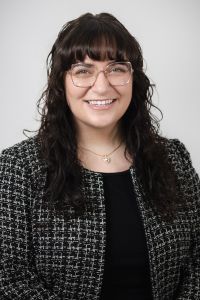 A professional headshot of a woman with glasses smiling, wearing a black top and textured blazer against a neutral background.