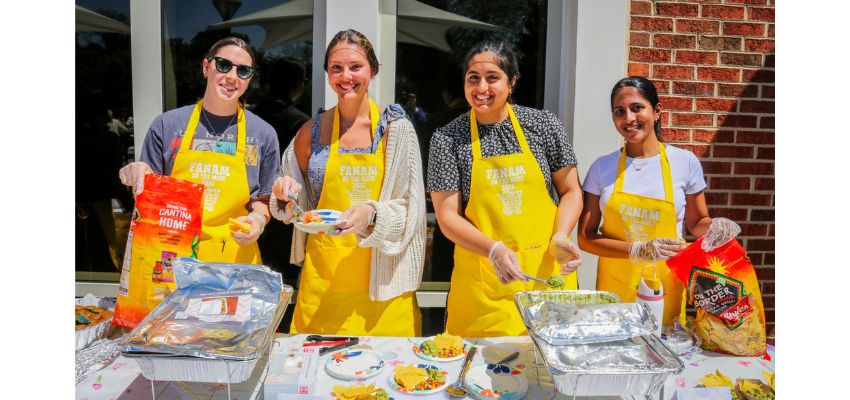 Four individuals wearing yellow aprons prepare and serve food outdoors at a table with trays and ingredients.