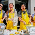 Fourindividualswearingyellowapronsprepareandservefoodoutdoorsatatablewithtraysandingredients