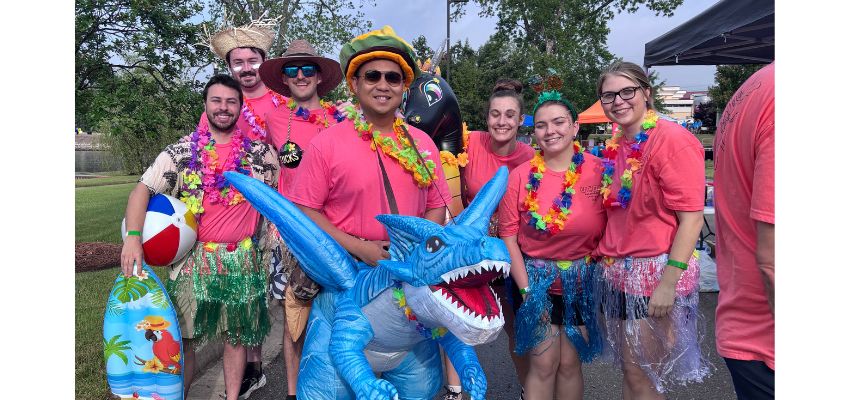 A group of people dressed in colorful, tropical-themed outfits pose together outdoors, some holding inflatable beach items.