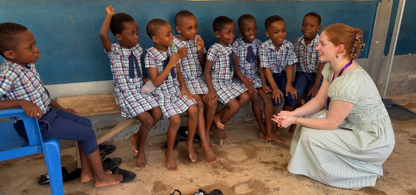 A woman sits and speaks with a group of young children in school uniforms seated on a bench in a classroom setting.