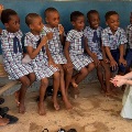 Awomansitsandspeakswithagroupofyoungchildreninschooluniformsseatedonabenchinaclassroomsetting