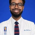 Professionalheadshotofastudentphysicianwearingawhitecoatglassesandstripedtieagainstabluebackground