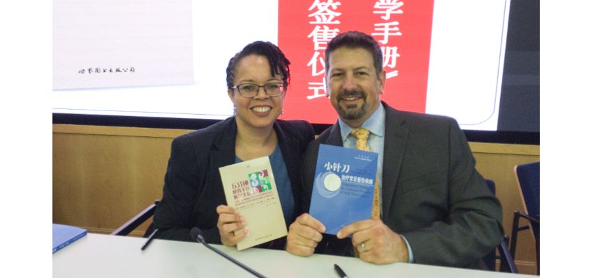Two individuals seated at a table hold books during an event featuring Chinese-language materials and presentation slides.