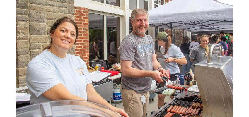 Two people grill food outdoors while others assist at a community cookout event.