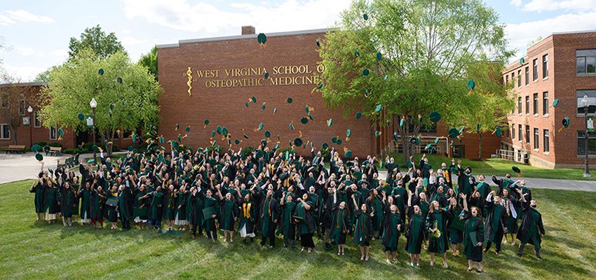 Graduates in caps and gowns toss their hats in the air outside the West Virginia School of Osteopathic Medicine building.