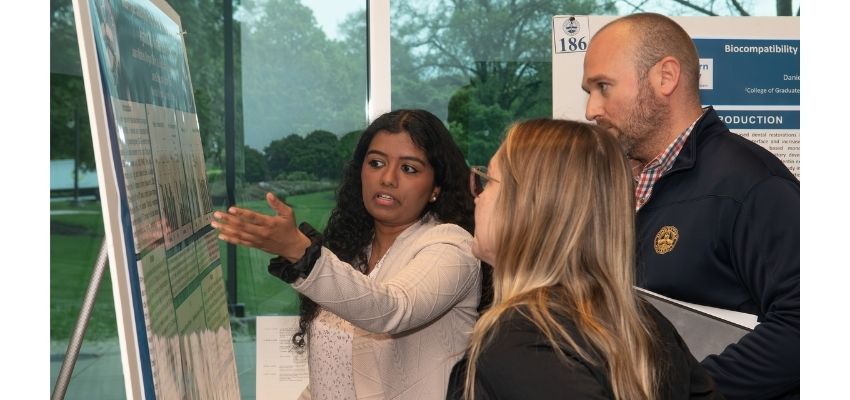 A student presents research findings on a poster while speaking with attendees at a poster session.