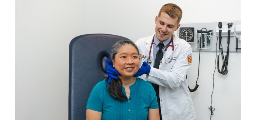A student physician examines a seated patient’s neck in a clinical exam room.