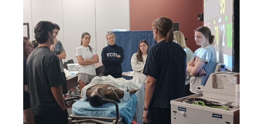 A group of students participates in a medical simulation training session around a patient mannequin in a classroom.