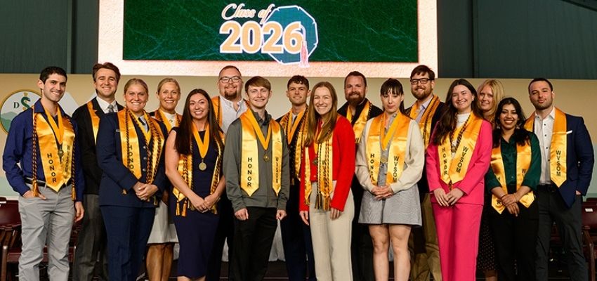 A group of students wearing honor cords and medallions poses on stage beneath a “Class of 2026” sign.