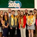 AgroupofstudentswearinghonorcordsandmedallionsposesonstagebeneathaClassof2026sign