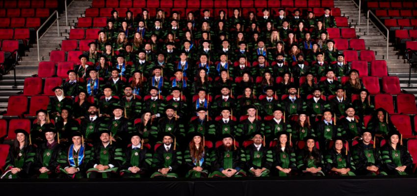 A large group of graduates in academic regalia poses together in an arena with red stadium seating in the background.