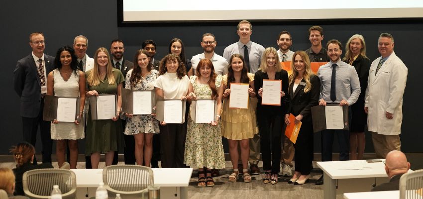 Students and faculty pose together indoors while holding certificates and awards during a recognition ceremony.