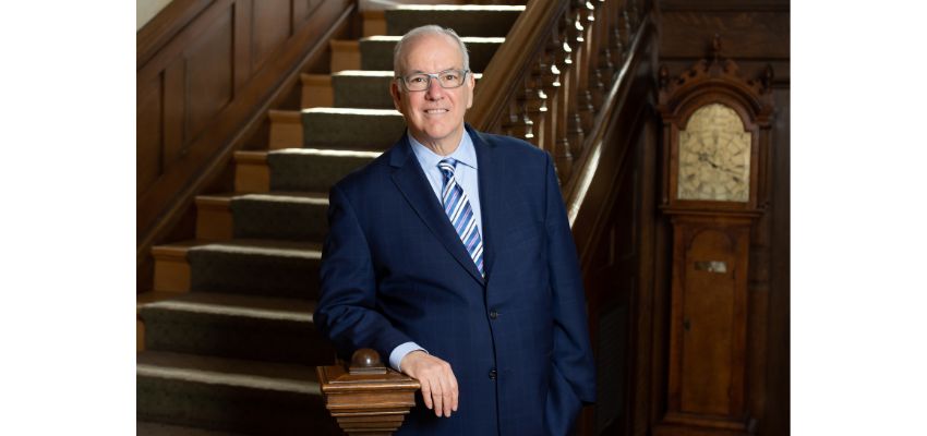 A man in a blue suit and striped tie stands beside a wooden staircase and grandfather clock in a formal interior setting.