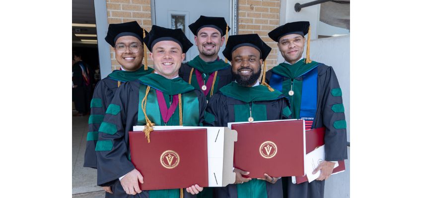 Five graduates in academic regalia smile and hold diploma covers outdoors following a graduation ceremony.