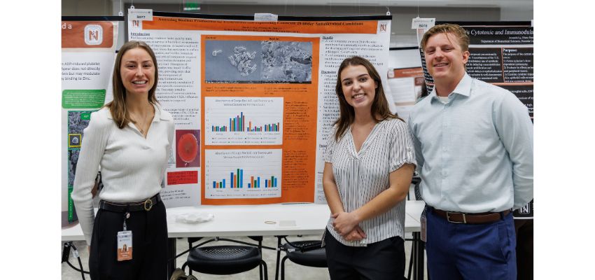 Three students stand in front of a research poster and smile during a research symposium or poster session.