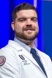 Professional headshot of a student physician wearing a white coat and tie against a blue background.