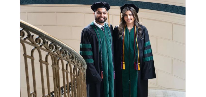 Two graduates in academic regalia pose together beside a decorative staircase inside a historic building.